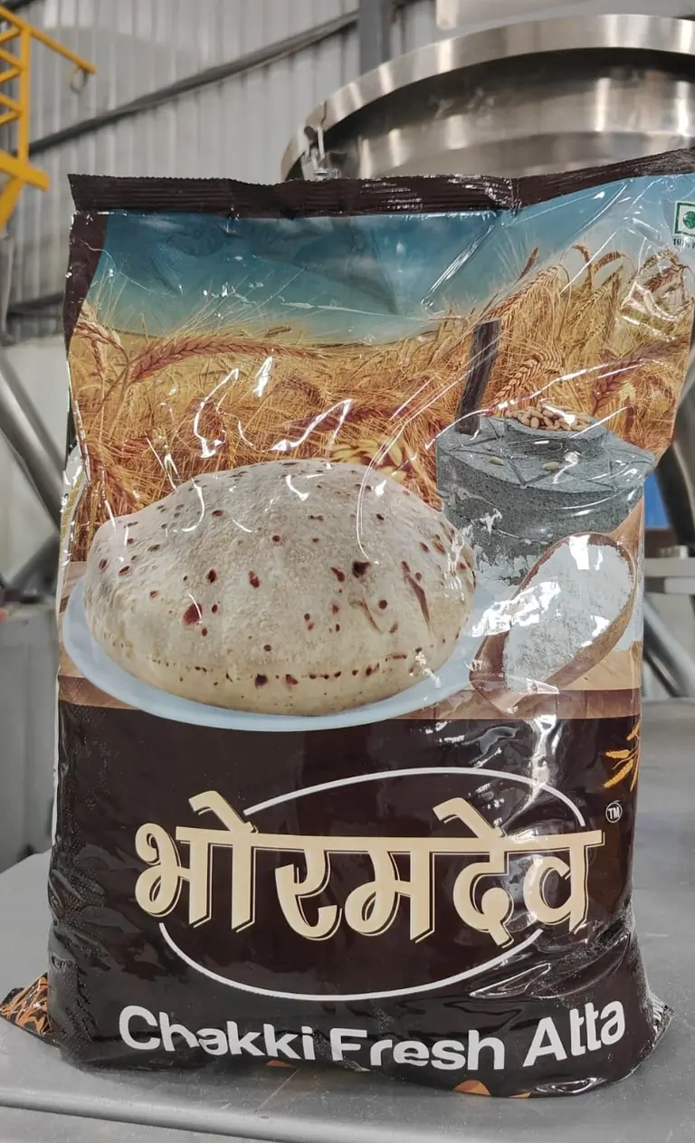 A 5 kg pack of Bhoramdev Chakki Fresh Atta on the packing floor, with a steel hopper behind it